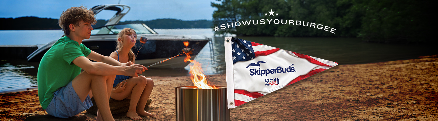 Two people sit by a lakeside campfire, roasting marshmallows. An American flag-themed SkipperBud's flag waves nearby.