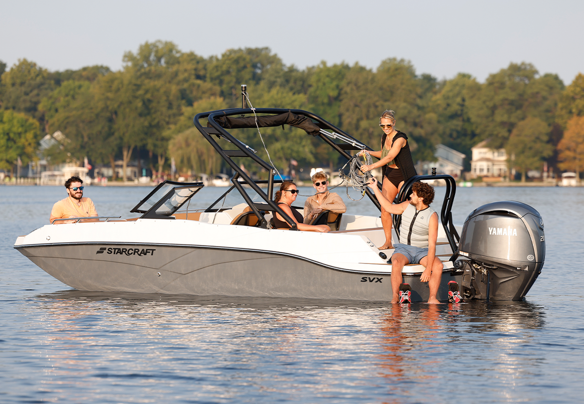 A Starcraft boat on a lake with several people on and around the boat, including one standing at the helm and two sitting with their feet in the water. Trees and houses are visible in the background.